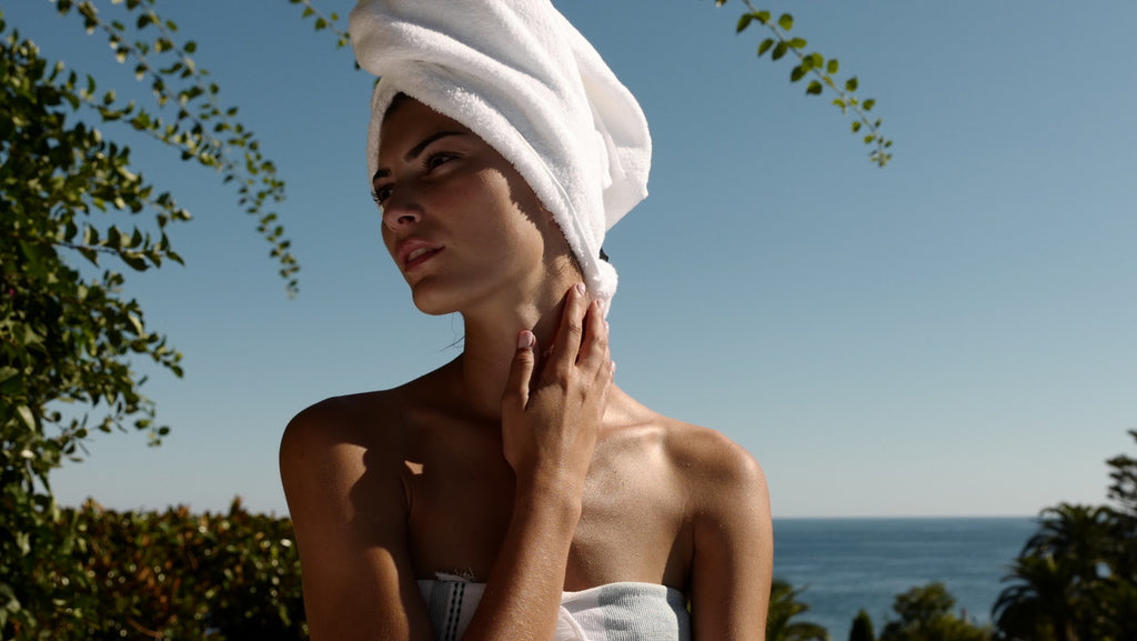 woman in a white towel and with white towel wrapped around her head, looking dreamy against a blue sky, sea and trees