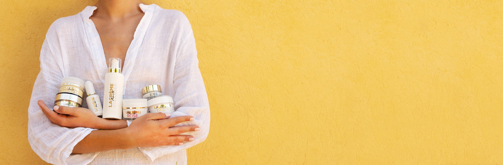 woman wearing a white linen shirt, holding a range of lacreme beaute skincare products in her arms, against a sunny yellow wall